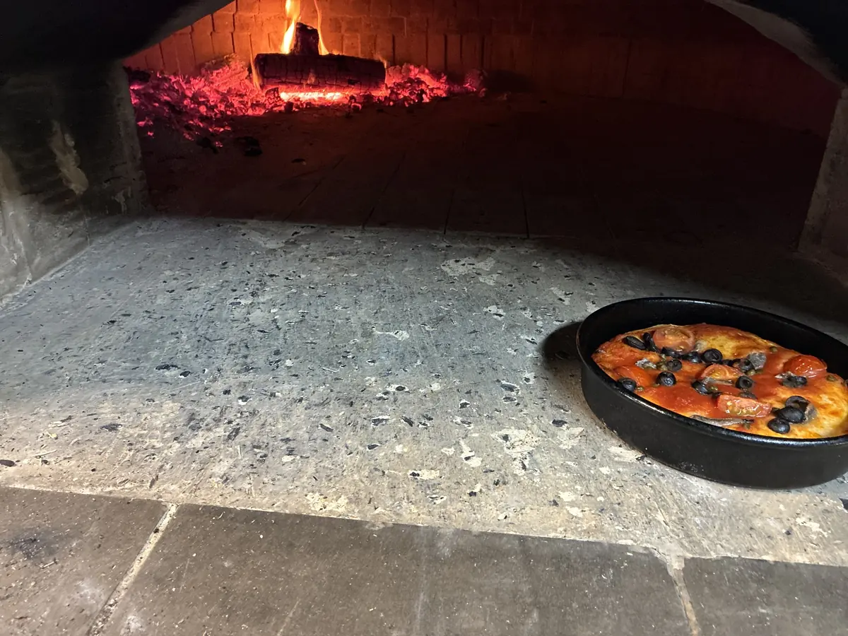 A pizza baking on the stone floor of the Tano’s wood-fired oven, with live coals and flame at the back