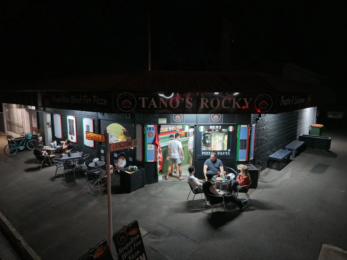 Drone photo of the Tano’s Rocky shopfront at night, glowing sign and tables out the front