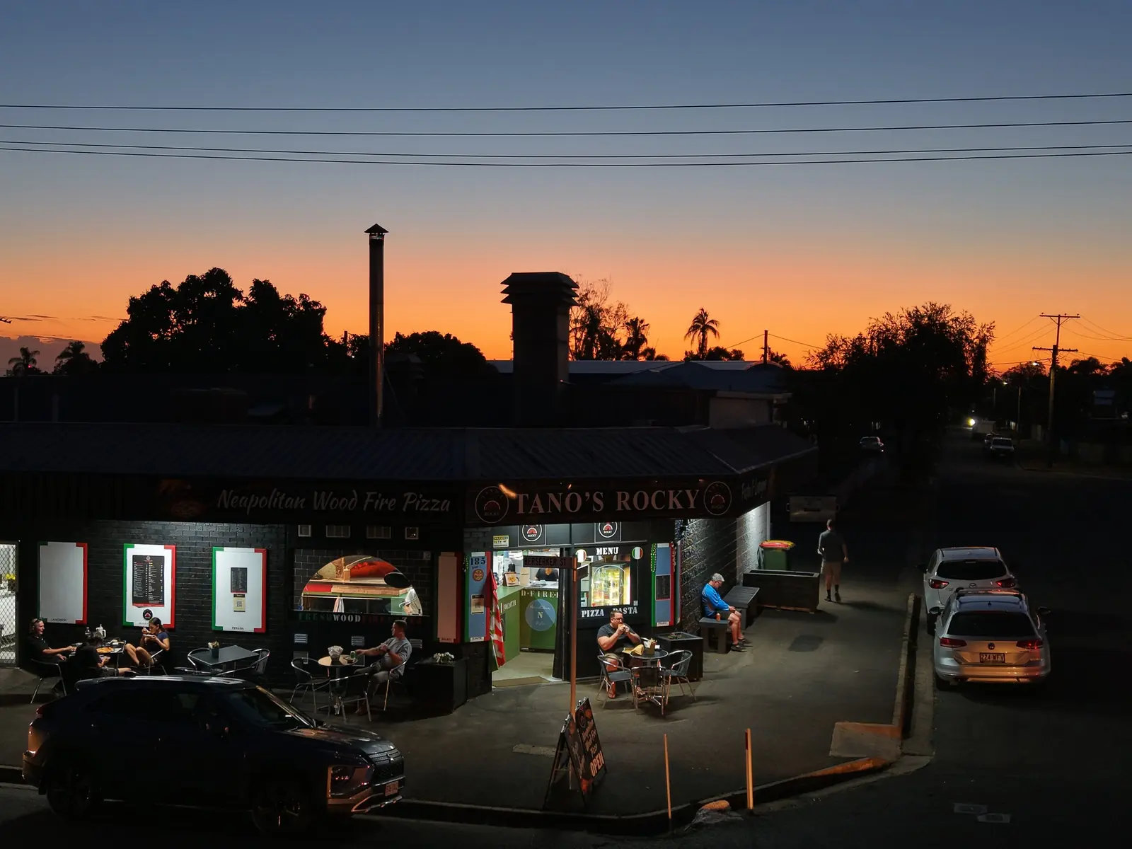 Drone photo of Tano’s Rocky at sunset, lit shopfront and outdoor tables under an orange sky